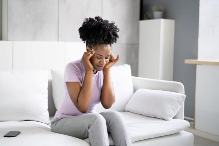 Woman on a couch in a modern living room with her hands against her temples and her head turned downwards. Her facial expression is neutral, but her eyes are closed. 